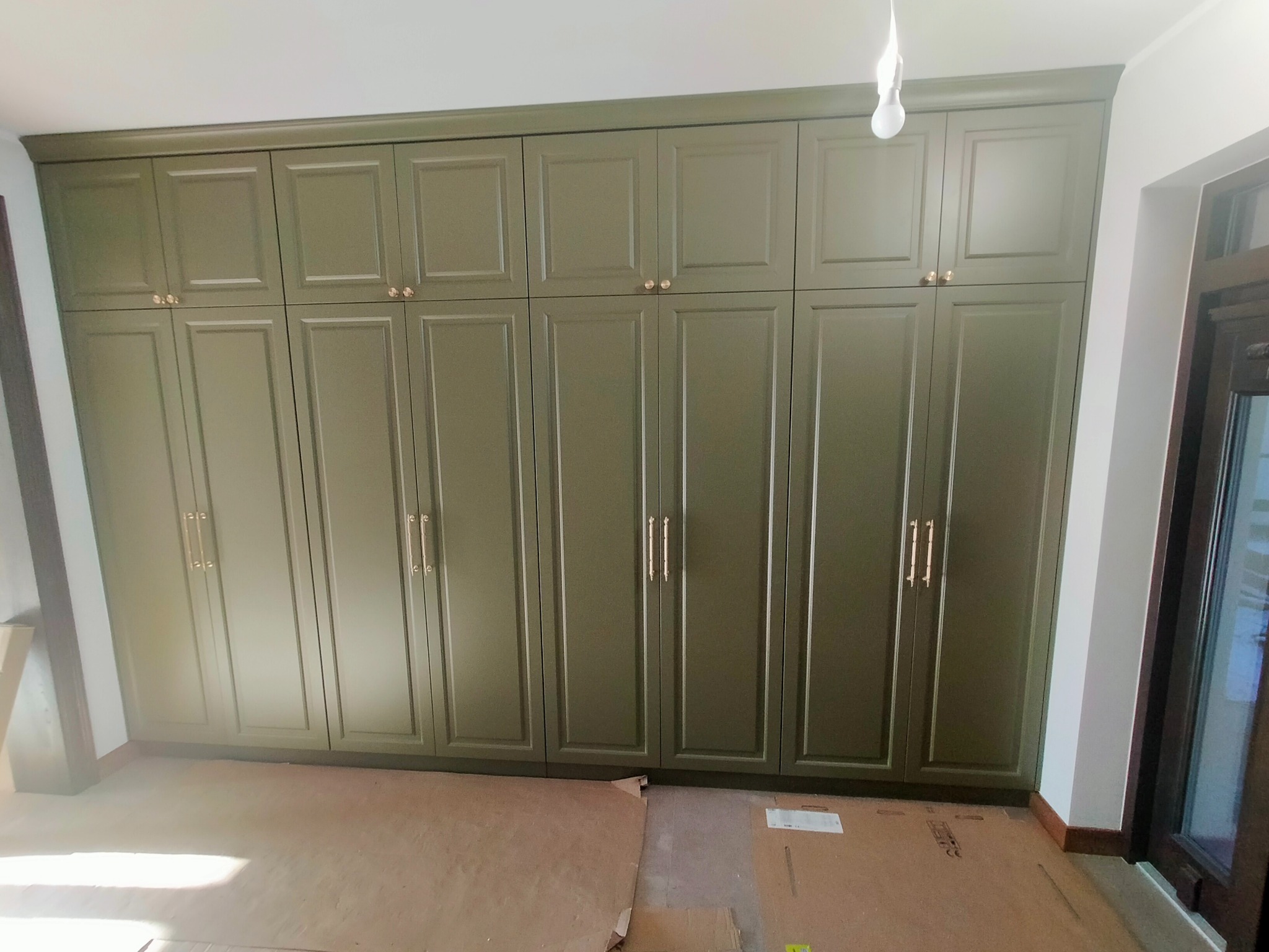 Duża, oliwkowa szafa wnękowa z frezowanymi frontami i złotymi uchwytami, zajmująca całą ścianę, na podłodze kartony zabezpieczające, widoczna żarówka.
