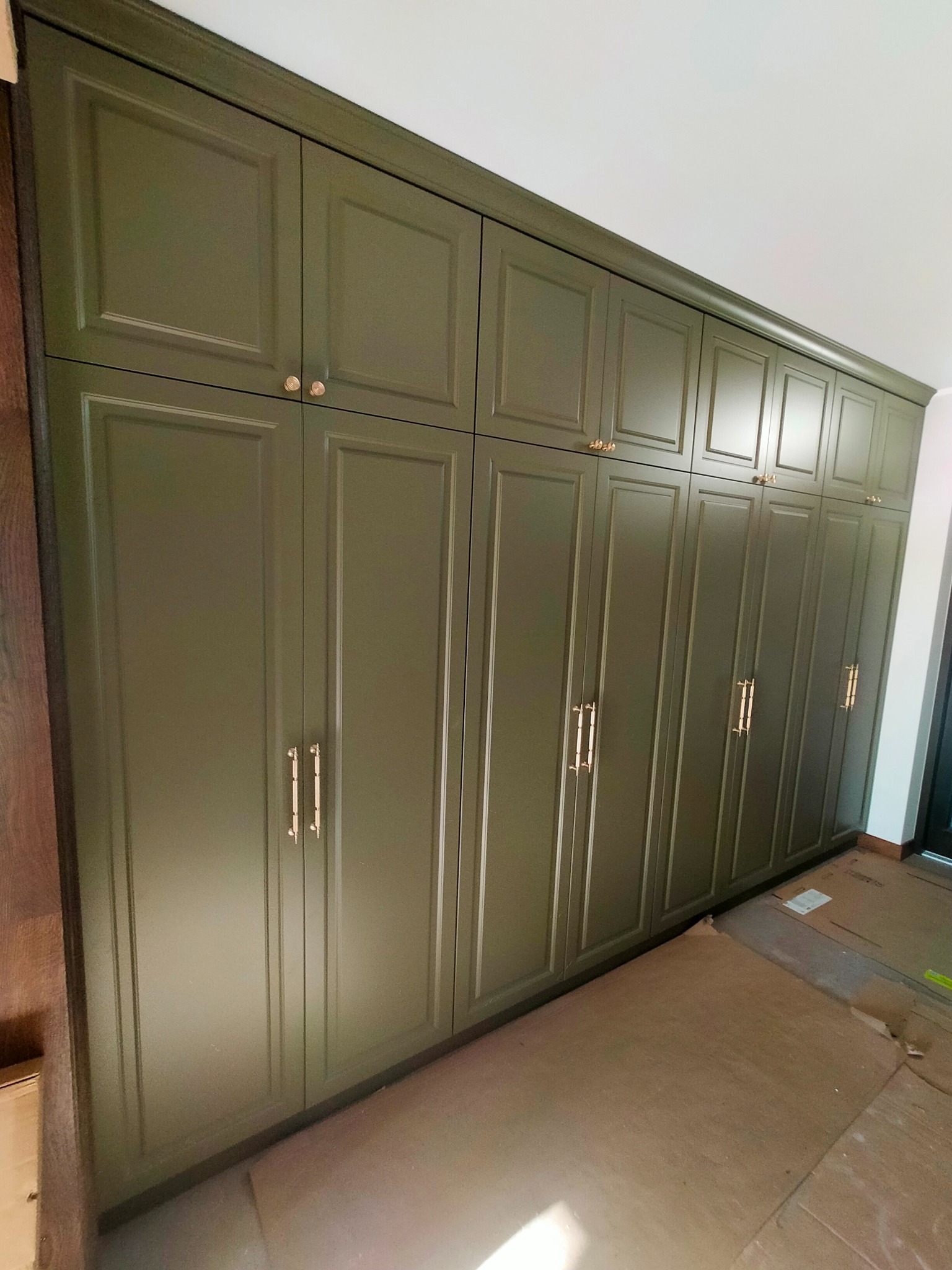 Długa, oliwkowa szafa wnękowa z frezowanymi drzwiami i złotymi uchwytami, sięgająca sufitu. Podłoga zabezpieczona kartonem.