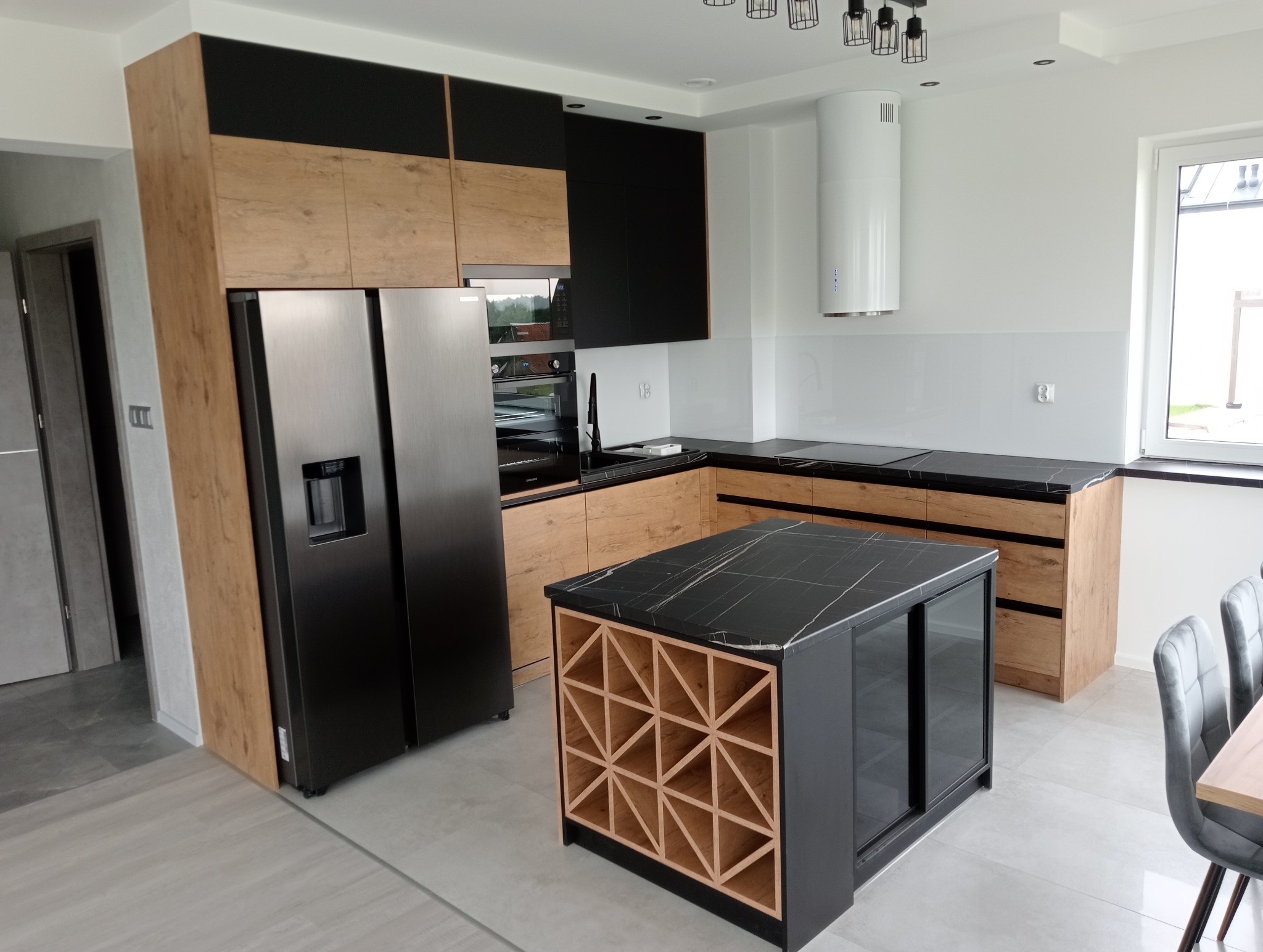 Nowoczesna kuchnia z wyspą: drewniane i czarne szafki, marmurowy blat, srebrna lodówka side-by-side, biały okap cylindryczny, szare krzesła.