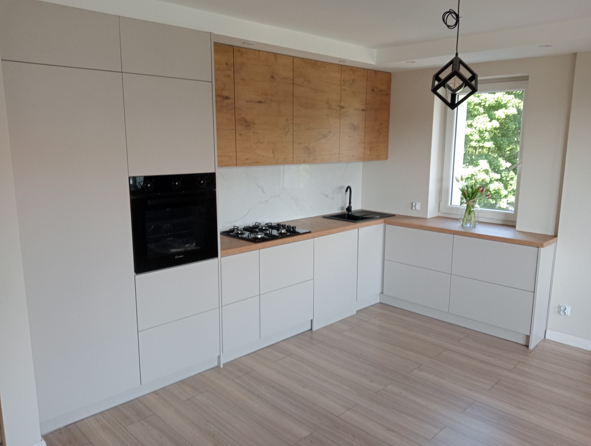 Nowoczesna kuchnia z białymi szafkami i drewnianymi akcentami. Czarny piekarnik w zabudowie, marmurowy backsplash i designerska lampa. Widok na okno z zielenią.