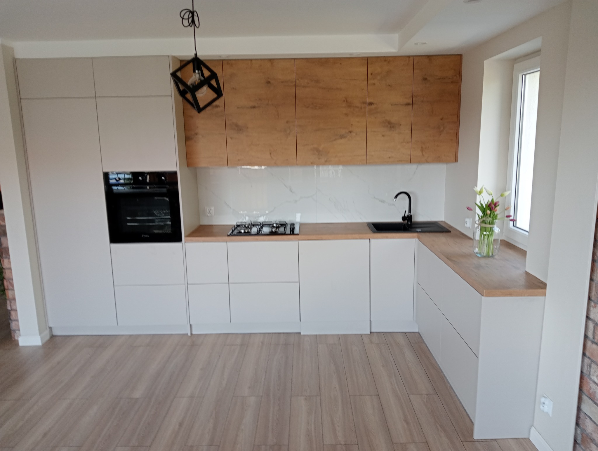 Nowoczesna kuchnia z białymi szafkami dolnymi i drewnopodobnymi górnymi, czarnym piekarnikiem i geometryczną lampą. Blat drewniany, marmurowe wykończenie ściany.
