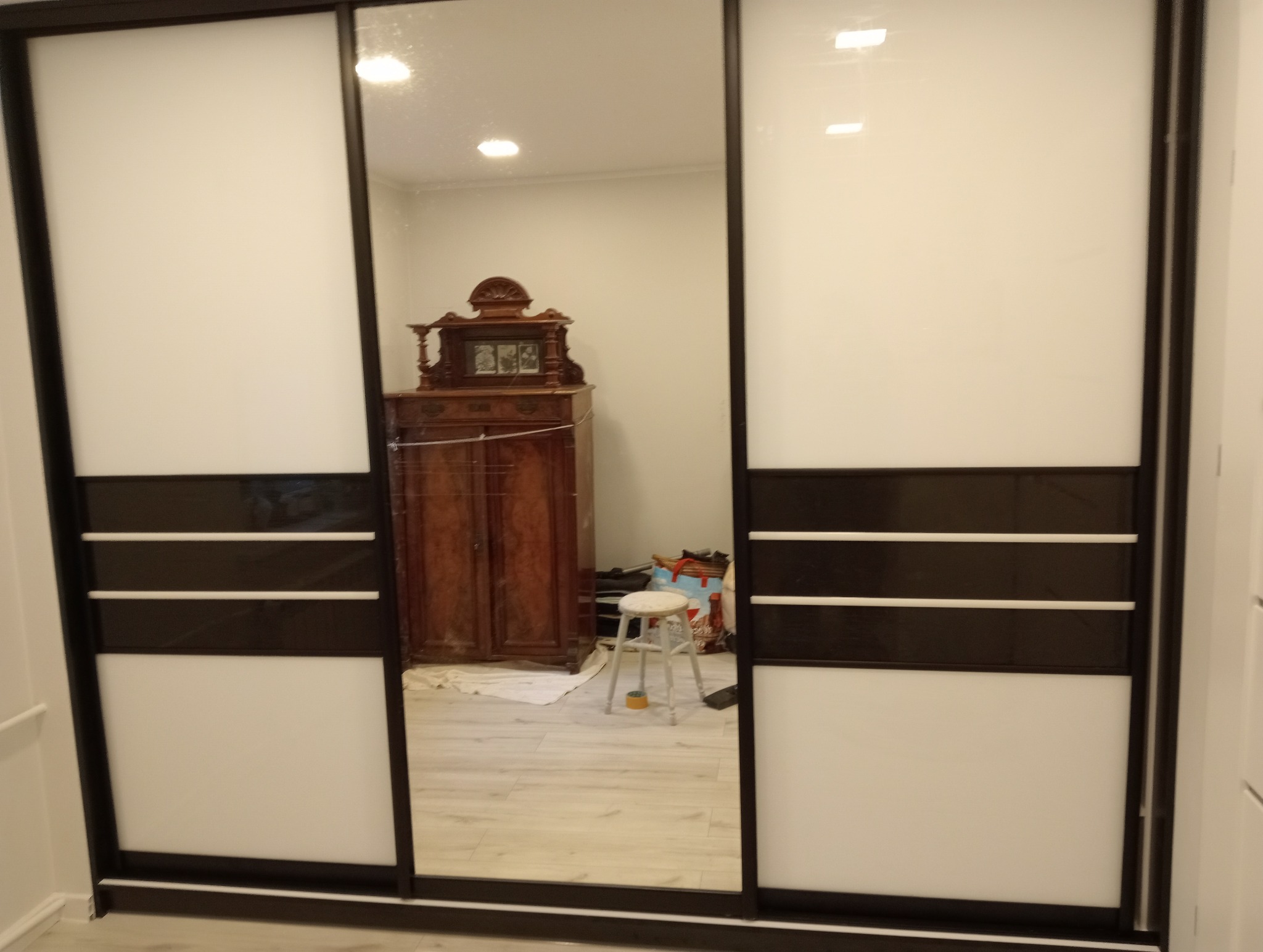 Nowoczesna szafa wnękowa z przesuwnymi drzwiami, dzielonymi poziomymi pasami w kolorze białym i ciemnobrązowym. W lustrzanym odbiciu widoczny antyczny kredens.