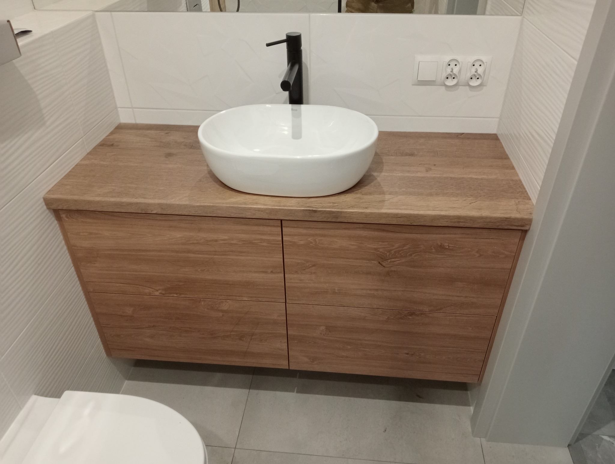 Drewniana szafka łazienkowa z owalną umywalką nablatową i czarną armaturą. Białe płytki z subtelnym wzorem na ścianie. Nowoczesny design.