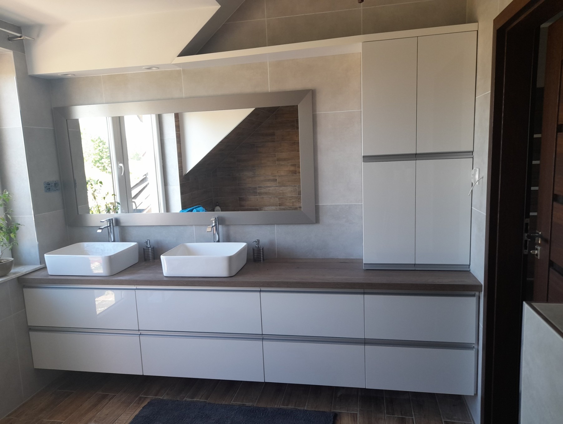 Nowoczesna łazienka w jasnych barwach: białe szafki pod umywalki nablatowe, duże lustro w szarej ramie, drewniany blat i płytki imitujące kamień. Minimalistyczny design.