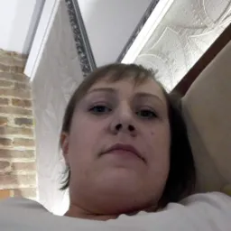 Kobieta z krótkimi włosami i niebieskimi oczami patrzy w dół na obiektyw, w tle widoczna ściana z cegły i wzorzysta tapeta.