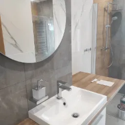 Nowoczesna łazienka po renowacji z okrągłym lustrem, białą umywalką nablatową na drewnianym blacie, szarą glazurą i kabiną prysznicową w tle.