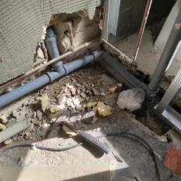 Szare rury kanalizacyjne łączone kształtkami w wykuciu w ścianie z resztkami płytek, widoczne fragmenty instalacji miedzianej, gruz, młotek i notatki na szkle.