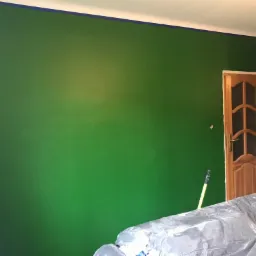 Świeżo pomalowana na zielono ściana z niebieską taśmą malarską przy suficie, drewniane drzwi po prawej stronie, meble zabezpieczone folią malarską na pierwszym planie, widoczny fragment wałka...