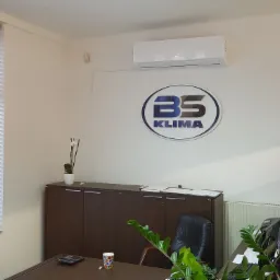 Biuro z zamontowaną klimatyzacją ścienną, logo firmy BS Klima na ścianie, biurko z roślinami i oknem z żaluzjami.