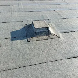 Płaski dach pokryty papą z widocznymi łączeniami i elementami wentylacyjnymi, jeden z nich z metalowym zabezpieczeniem.