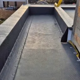 Szczegółowe ujęcie płaskiego dachu pokrytego hydroizolacją bitumiczną, z widoczną obróbką krawędzi i systemem odprowadzania wody, tło stanowi zachód słońca nad zabudowaniami.