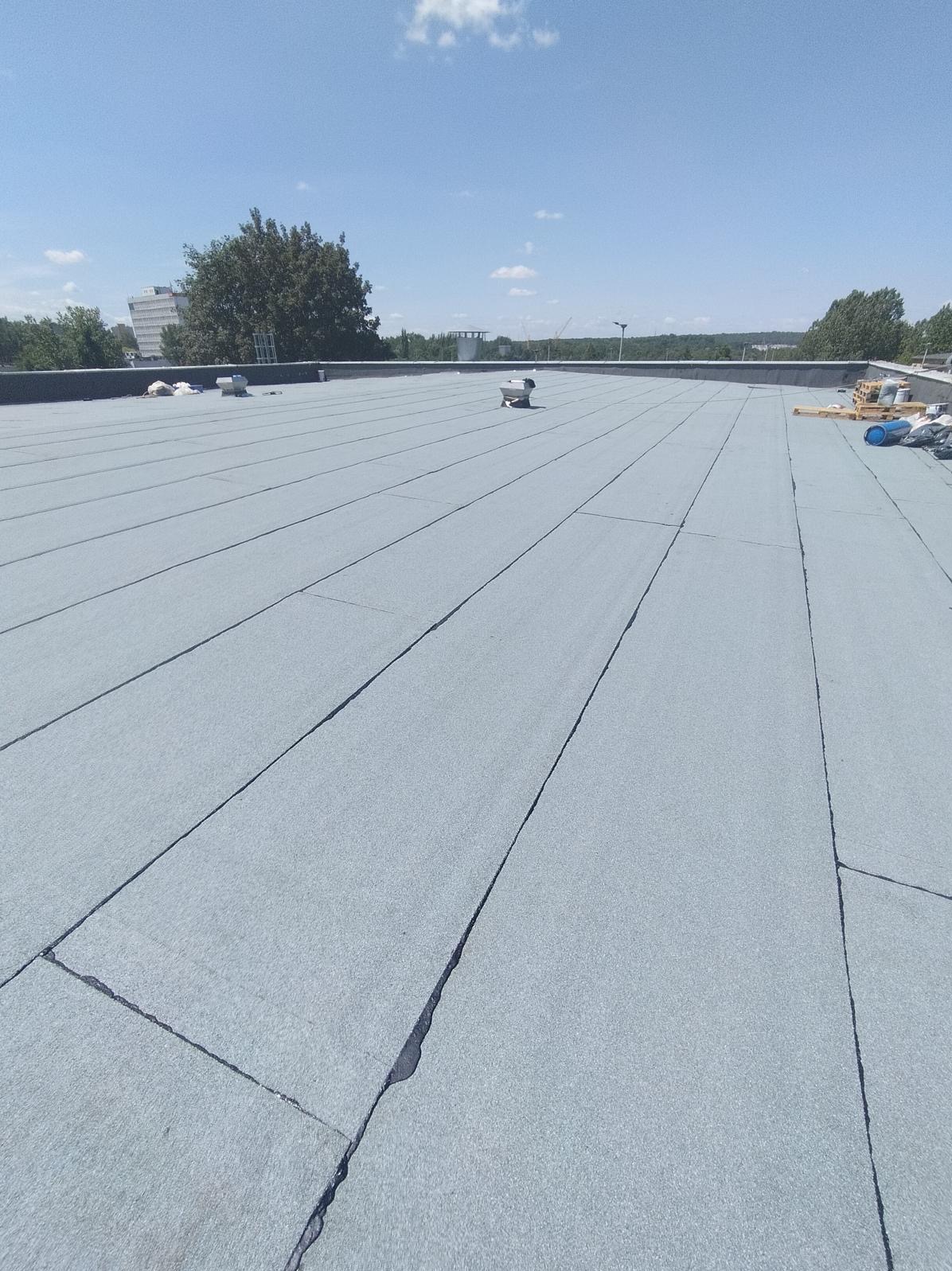 Widok z góry na płaski dach pokryty szarą papą termozgrzewalną, z widocznymi łączeniami i kominkami wentylacyjnymi, na horyzoncie drzewa i budynki pod błękitnym niebem.