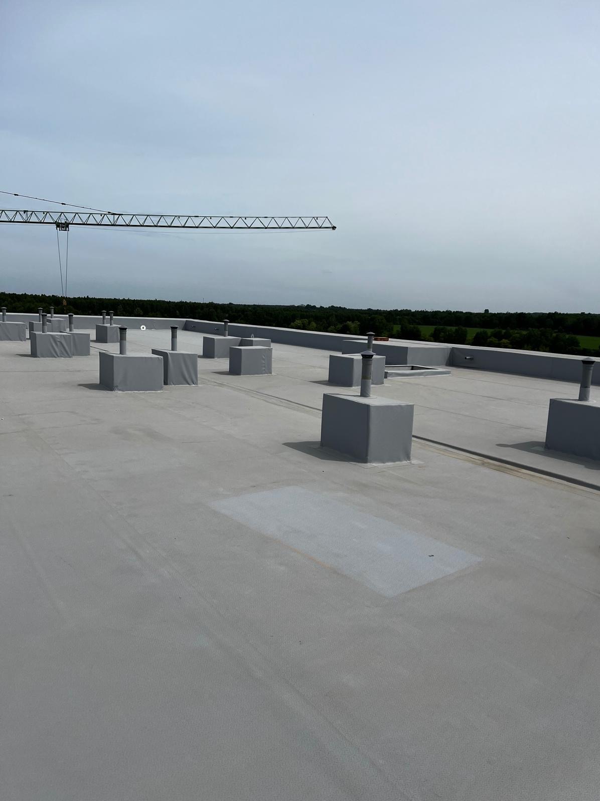 Widok z góry na płaski dach pokryty szarą membraną, z kominami wentylacyjnymi zabezpieczonymi szarymi obudowami, w tle widoczny las i fragment żurawia budowlanego.