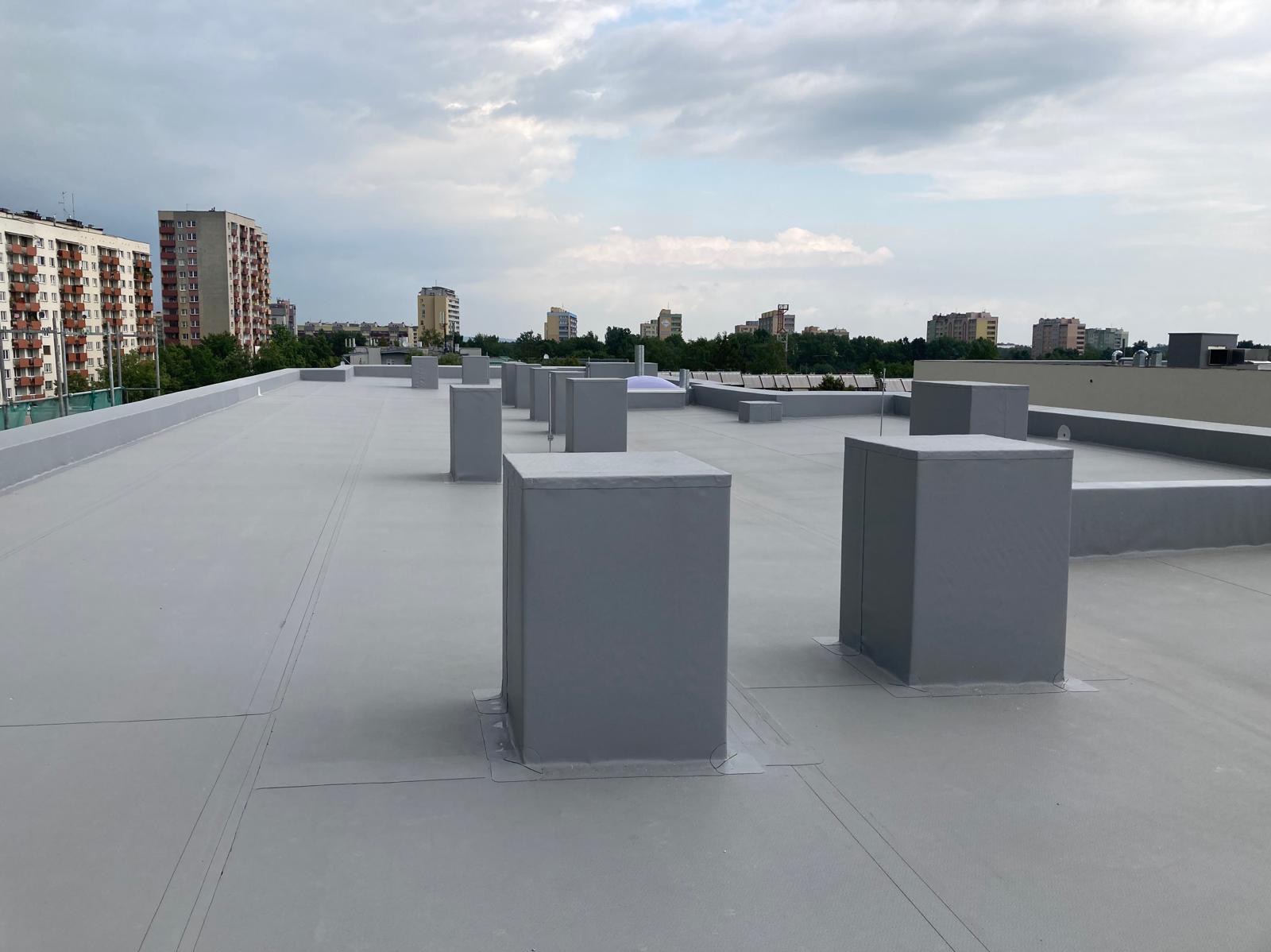 Szary, płaski dach pokryty papą termozgrzewalną z prostokątnymi obudowami wentylacyjnymi na tle budynków mieszkalnych i pochmurnego nieba.