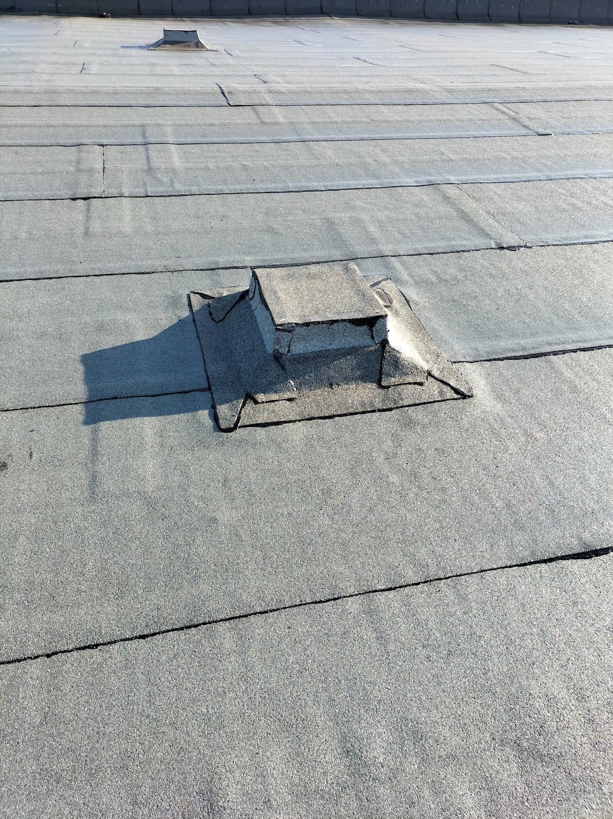 Płaski dach pokryty papą z widocznymi łączeniami i elementami wentylacyjnymi, jeden z nich z metalowym zabezpieczeniem.