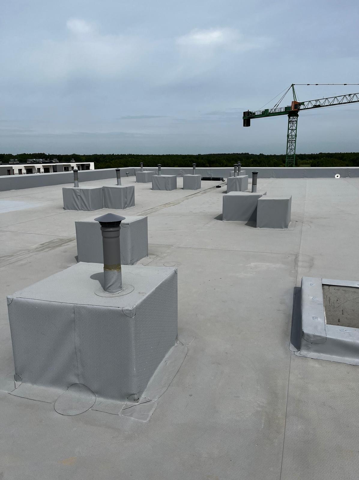 Szary płaski dach z licznymi kominami wentylacyjnymi w obudowach, widoczny żuraw budowlany w tle.