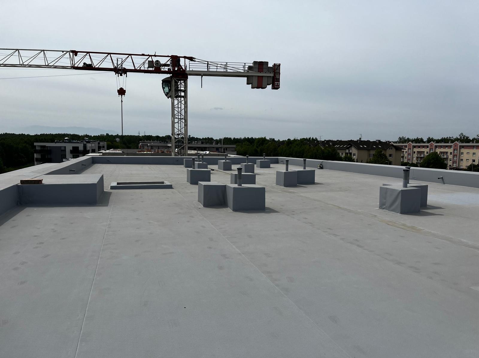 Szary, płaski dach budynku wielorodzinnego z widocznymi kominami wentylacyjnymi i attyką, w tle żuraw budowlany i zabudowa miejska