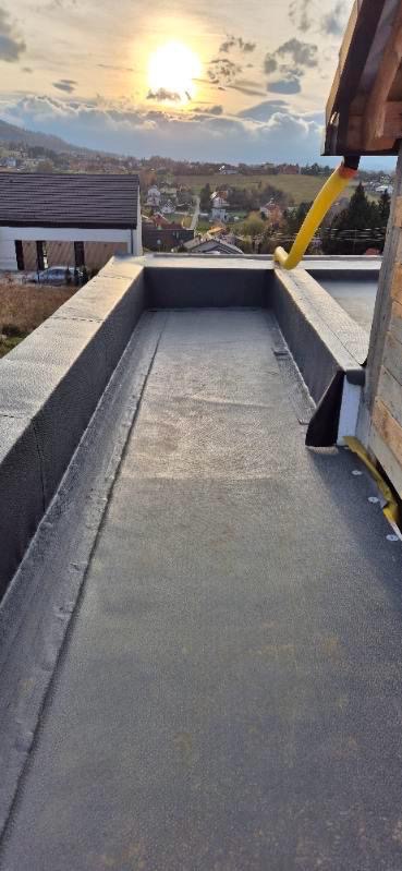 Szczegółowe ujęcie płaskiego dachu pokrytego hydroizolacją bitumiczną, z widoczną obróbką krawędzi i systemem odprowadzania wody, tło stanowi zachód słońca nad zabudowaniami.