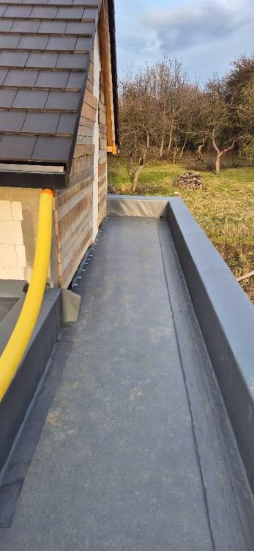 Widok z góry na płaski dach pokryty papą, z widoczną rynną i fragmentem ściany budynku z drewnianą elewacją oraz żółtą rurą spustową, w tle drzewa i trawa.