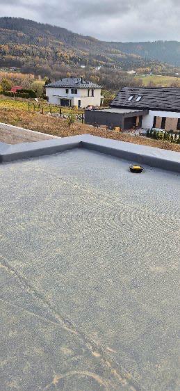 Szary, płaski dach pokryty papą termozgrzewalną z widocznym narzędziem dekarskim i panoramą górską w tle.