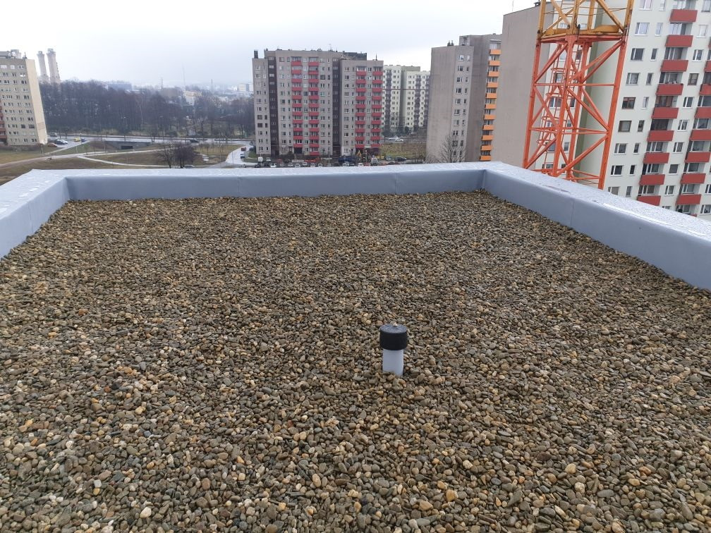 Widok z góry na płaski dach pokryty drobnymi kamieniami, z widocznym kominkiem wentylacyjnym i szarym obrzeżem, w tle bloki mieszkalne i żuraw budowlany w pochmurny dzień.