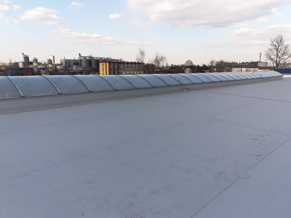 Płaski dach pokryty jasną membraną z zainstalowanymi świetlikami tunelowymi, w tle zabudowania przemysłowe i błękitne niebo z chmurami.
