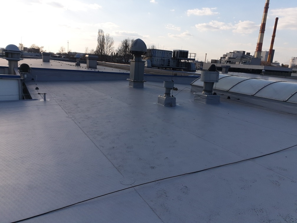 Widok z góry na płaski dach przemysłowy pokryty szarą membraną, z widocznymi kominami wentylacyjnymi i świetlikiem, w tle zabudowania przemysłowe i kominy elektrowni.