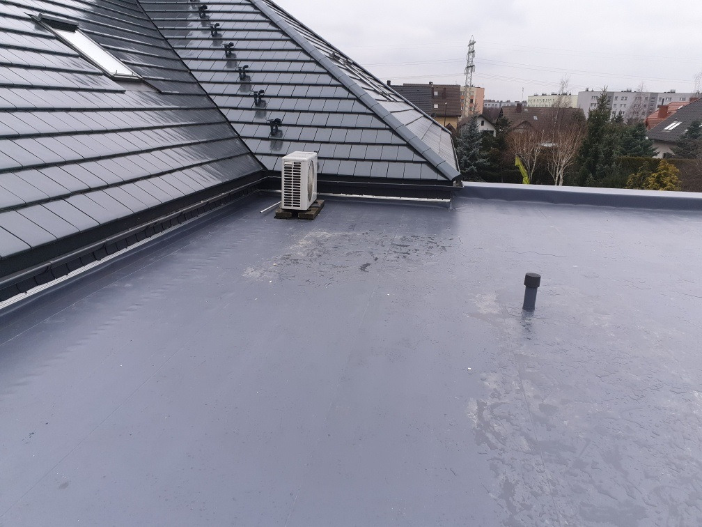 Szary, płaski dach pokryty hydroizolacją z widocznym kominem wentylacyjnym i jednostką zewnętrzną klimatyzacji na tle dachu skośnego z szarej dachówki ceramicznej i zabudowy miejskiej w pochmurny...