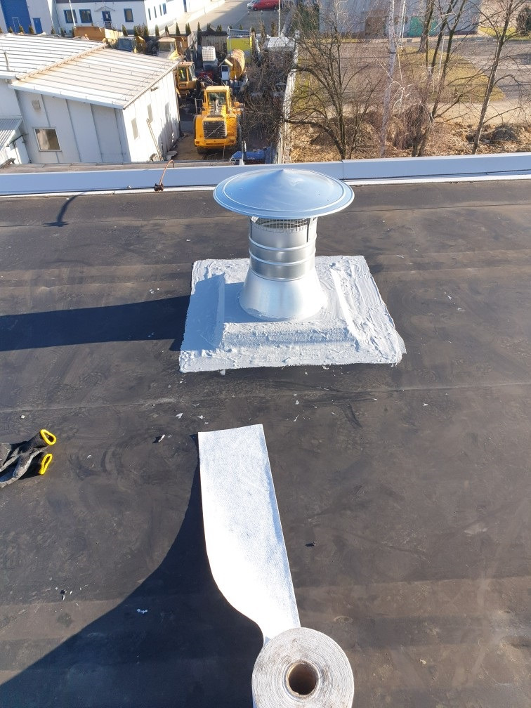 Metalowy komin wentylacyjny na czarnym, płaskim dachu, uszczelniony szarą masą, z rolką papy w dolnej części kadru i widocznymi rękawicami roboczymi, w tle budynki i sprzęt budowlany.