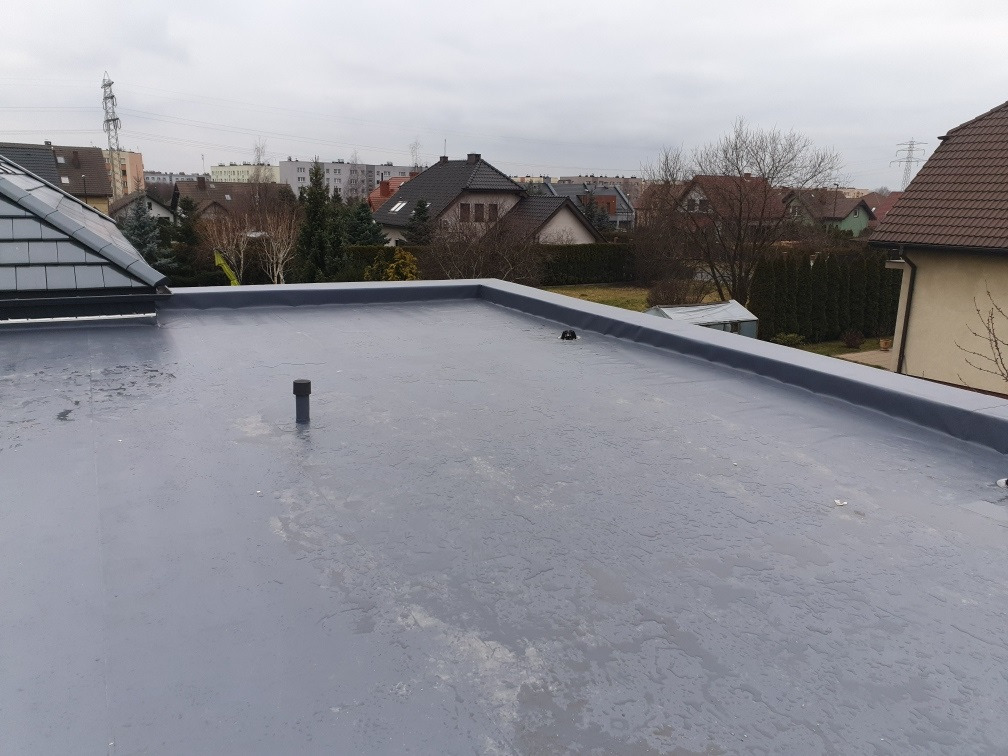 Widok z góry na płaski, szary dach z pojedynczym pionowym odpowietrznikiem, otoczony niskimi budynkami mieszkalnymi i pochmurnym niebem w tle.