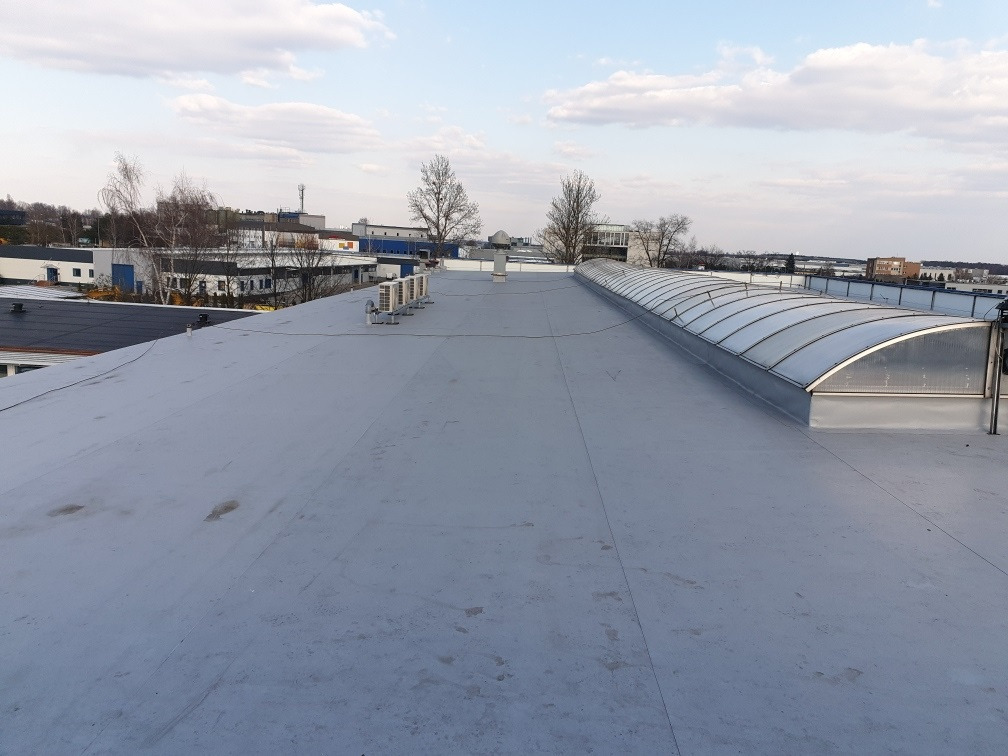 Widok z góry na szary, płaski dach przemysłowy z świetlikiem tunelowym i klimatyzatorami, w tle zabudowania przemysłowe i błękitne niebo z chmurami.