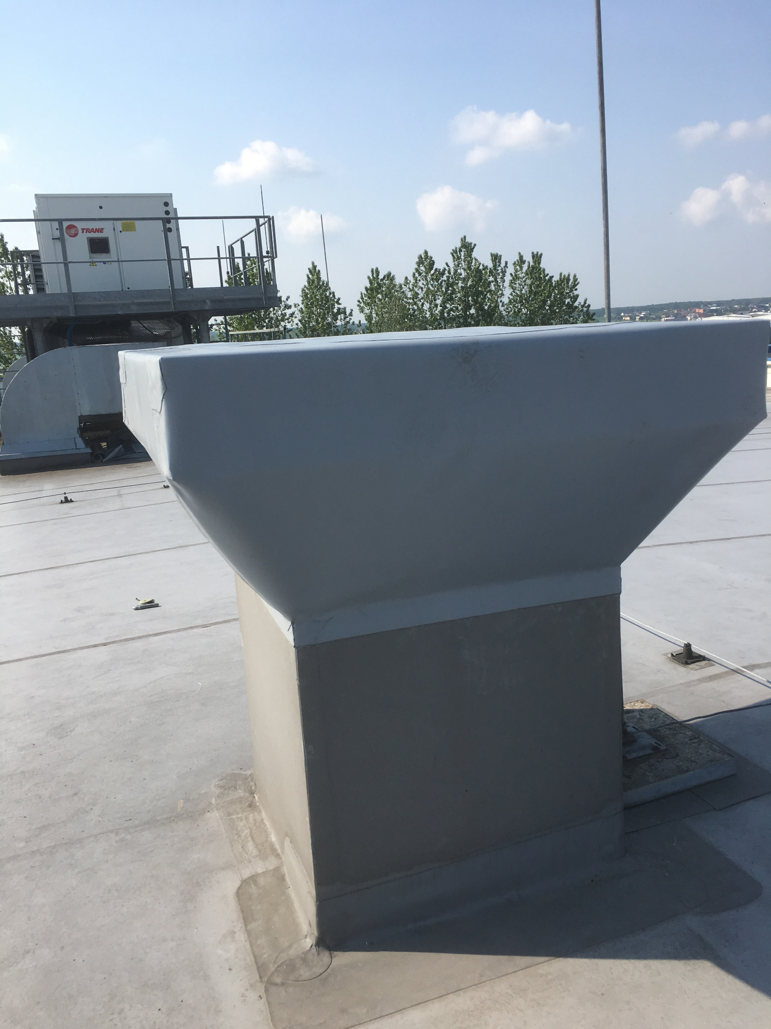 Szara obróbka blacharska komina wentylacyjnego na płaskim dachu, z widocznym systemem klimatyzacji Trane w tle i fragmentem panoramy miasta.