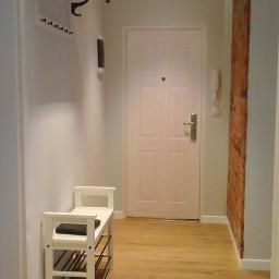 Pro Construction - Przedpokój w jasnych barwach z drewnianą podłogą, białą szafką na buty z czarnym siedziskiem, wieszakiem z półką i ceglanym fragmentem ściany po prawej stronie.