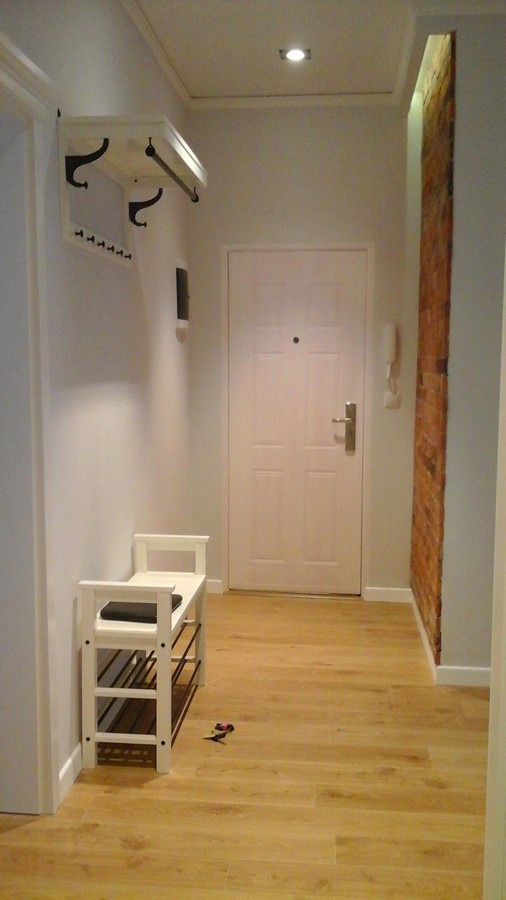 Przedpokój w jasnych barwach z drewnianą podłogą, białą szafką na buty z czarnym siedziskiem, wieszakiem z półką i ceglanym fragmentem ściany po prawej stronie.