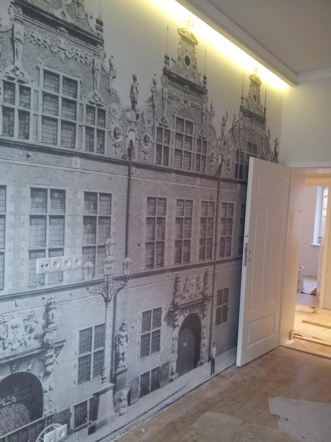 Fototapeta przedstawiająca kamienice w stylu historycznym na ścianie w pokoju, widoczne gniazdka elektryczne, oświetlenie LED podsufitowe i otwarte drzwi do kolejnego pomieszczenia z widocznym...