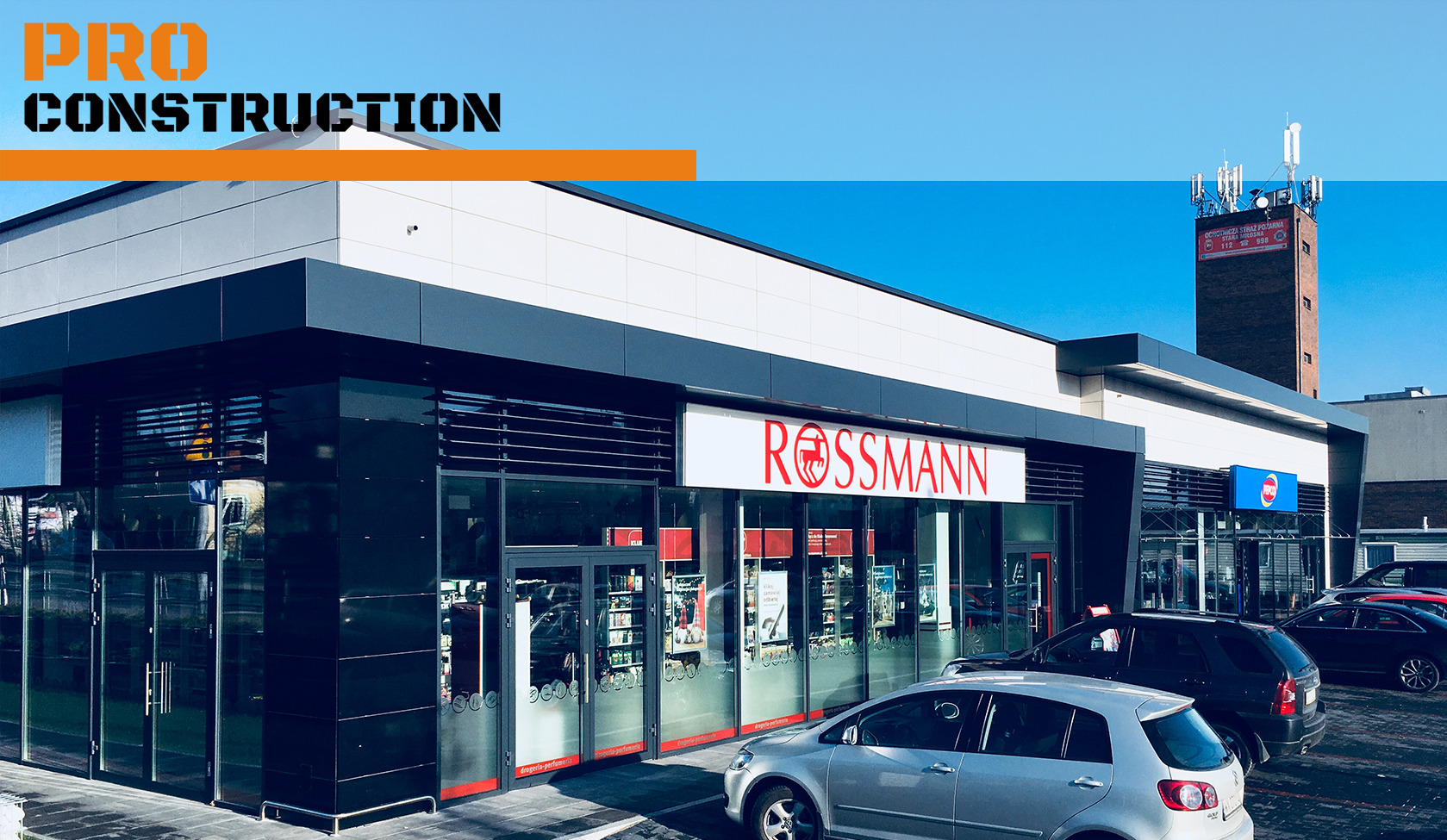 Nowoczesny budynek sklepu Rossmann z czarnymi panelami elewacyjnymi i dużymi witrynami, widoczny z zewnątrz na tle błękitnego nieba, z zaparkowanymi samochodami przed wejściem.
