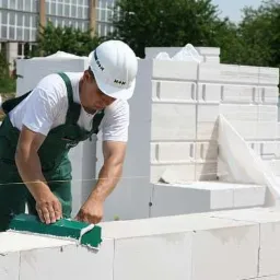 Murarz w zielonym kombinezonie wyrównuje bloczki z betonu komórkowego na wznoszonej ścianie, używając zielonej poziomicy, z widoczną konstrukcją budynku w tle.