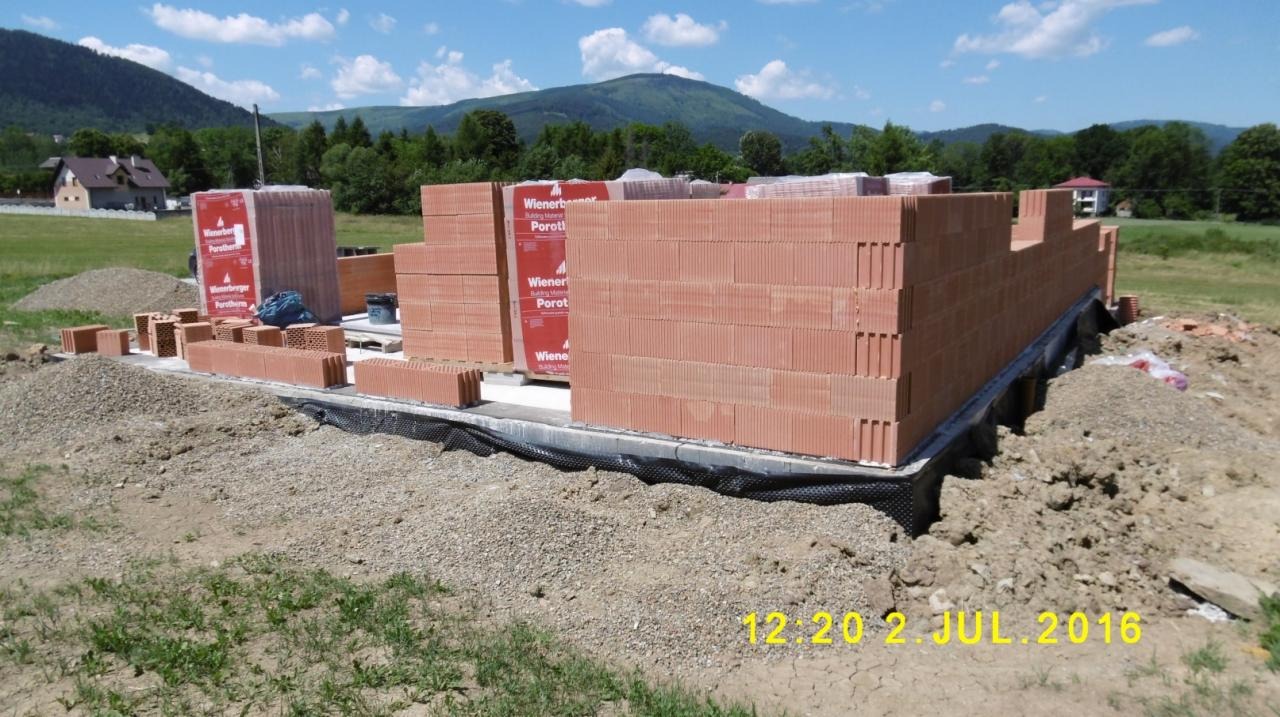 Wznoszenie ścian domu z czerwonej ceramiki Porotherm na fundamencie z izolacją przeciwwilgociową, w tle górzysty krajobraz i częściowo widoczny budynek mieszkalny.