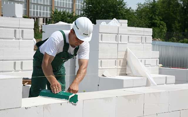 Murarz w zielonym kombinezonie wyrównuje bloczki z betonu komórkowego na wznoszonej ścianie, używając zielonej poziomicy, z widoczną konstrukcją budynku w tle.
