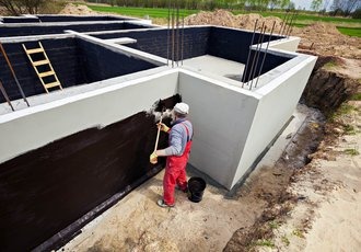 Pracownik w czerwonym kombinezonie maluje hydroizolacją betonowe ściany fundamentowe pędzlem. Widoczne stalowe pręty zbrojeniowe, drabina oparta o ścianę i wiadro z preparatem na piasku.