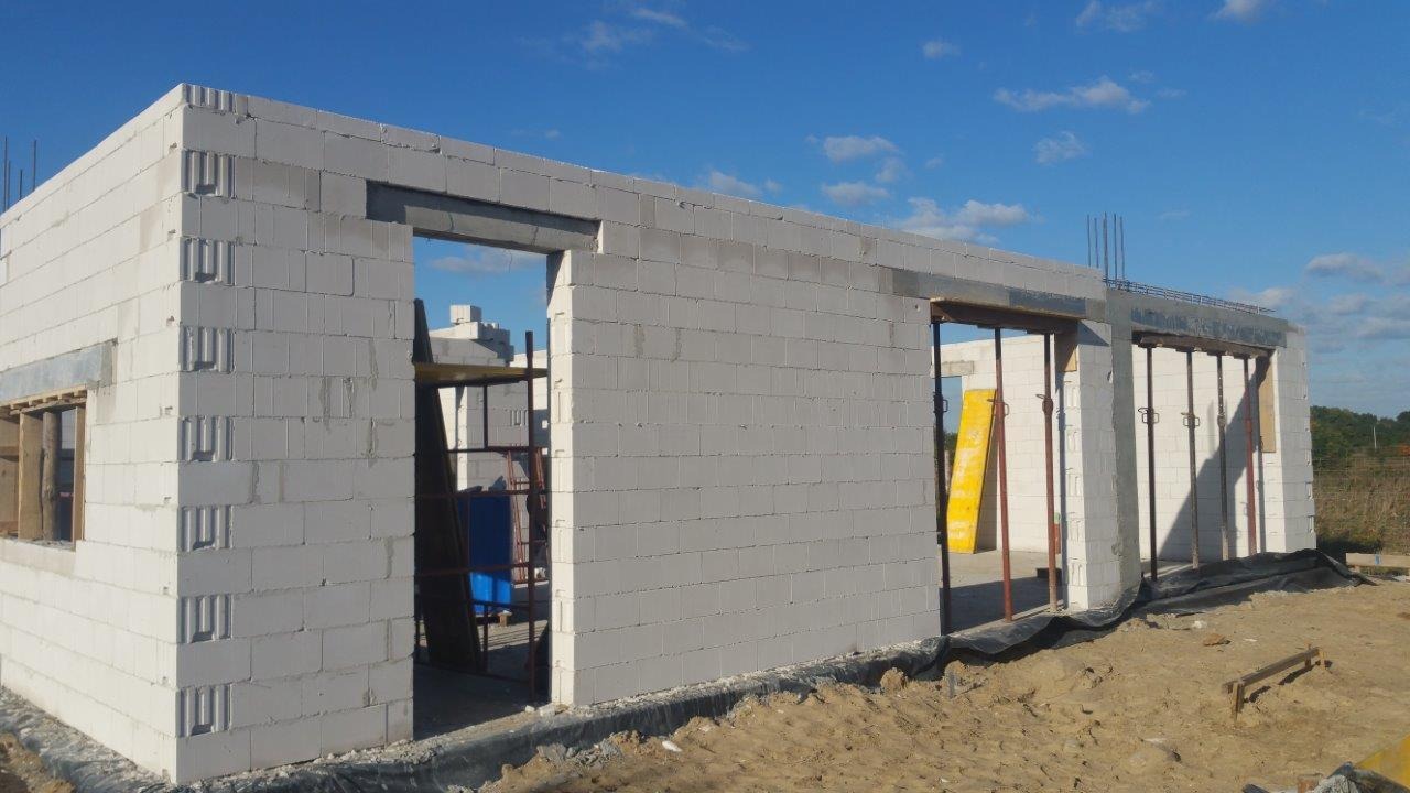 Wznoszenie ścian parterowego domu z białych bloczków betonu komórkowego, widoczne otwory okienne i drzwiowe z nadprożami, szalunki i zbrojenia w górnej części ścian.