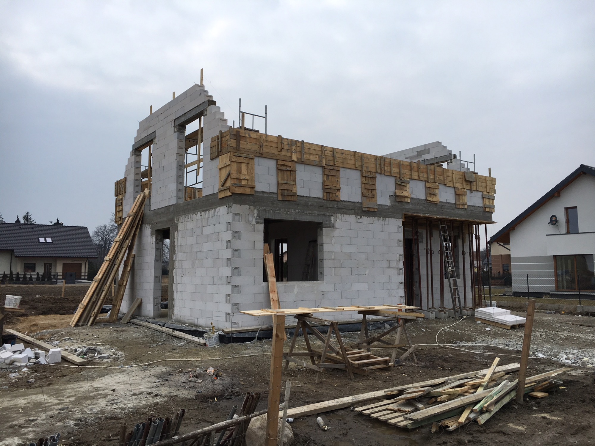 Niedokończony budynek z betonu komórkowego w trakcie murowania ścian, z widocznym drewnianym szalunkiem stropu i stertą desek na placu budowy, sąsiedni dom w tle.