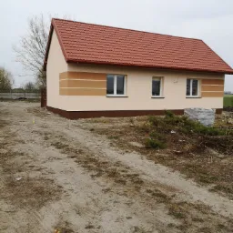 Jednorodzinny dom z nową, czerwoną dachówką i świeżo pomalowaną elewacją w odcieniach beżu i brązu, z widocznymi trzema oknami i stertą gałęzi na niezagospodarowanym terenie.