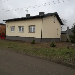 Jednopiętrowy dom z beżową elewacją, ciemną podmurówką i grafitowym dachem w pochmurny dzień, widoczne trzy okna z białymi ramami i małe drzewka iglaste przed budynkiem.