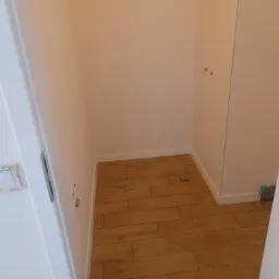 Wnęka z wykończonymi ścianami w odcieniu złamanej bieli i podłogą wyłożoną panelami imitującymi drewno, widoczne ślady po instalacjach elektrycznych i sanitarnych, listwy przypodłogowe.