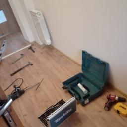Wykończenia wnętrz - Rozłożone narzędzia do montażu drzwi: wiertarka, młotki, poziomica, skrzynki narzędziowe Makita i DeWalt na drewnianej podłodze w pobliżu ściany i grzejnika.