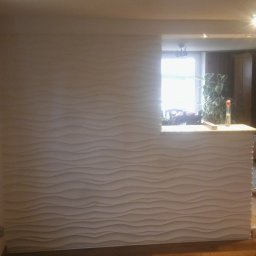 Wykończenia wnętrz - Ściana z dekoracyjną, falistą strukturą 3D, oddzielająca przestrzeń, z widocznym oknem i wyposażeniem wnętrza w tle.