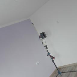 Wykończenia wnętrz - Wnętrze w trakcie prac wykończeniowych: widoczny statyw z narzędziem do szlifowania ścian pod kątem, ściana pomalowana częściowo na fioletowo, częściowo na biało, drewnopodobna podłoga oraz lampa...