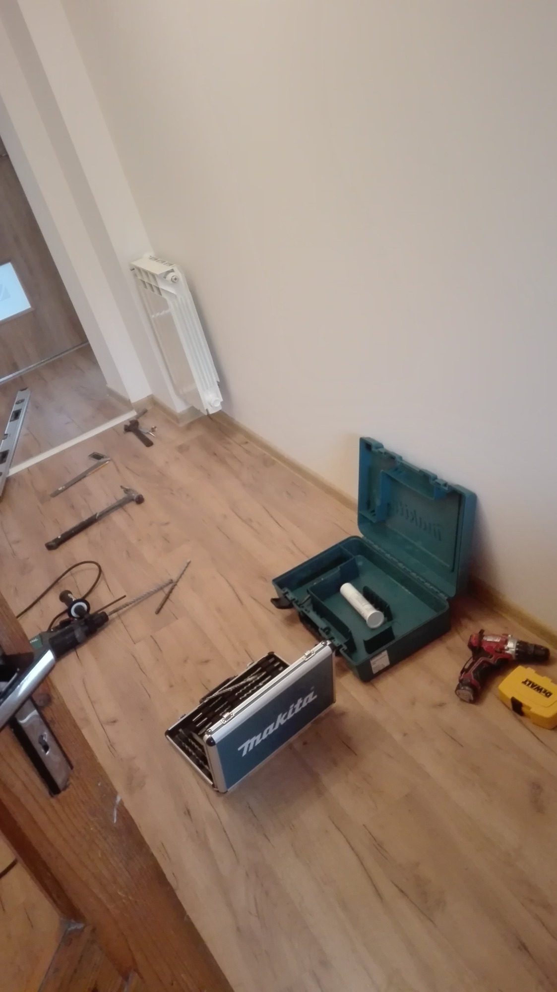 Rozłożone narzędzia do montażu drzwi: wiertarka, młotki, poziomica, skrzynki narzędziowe Makita i DeWalt na drewnianej podłodze w pobliżu ściany i grzejnika.