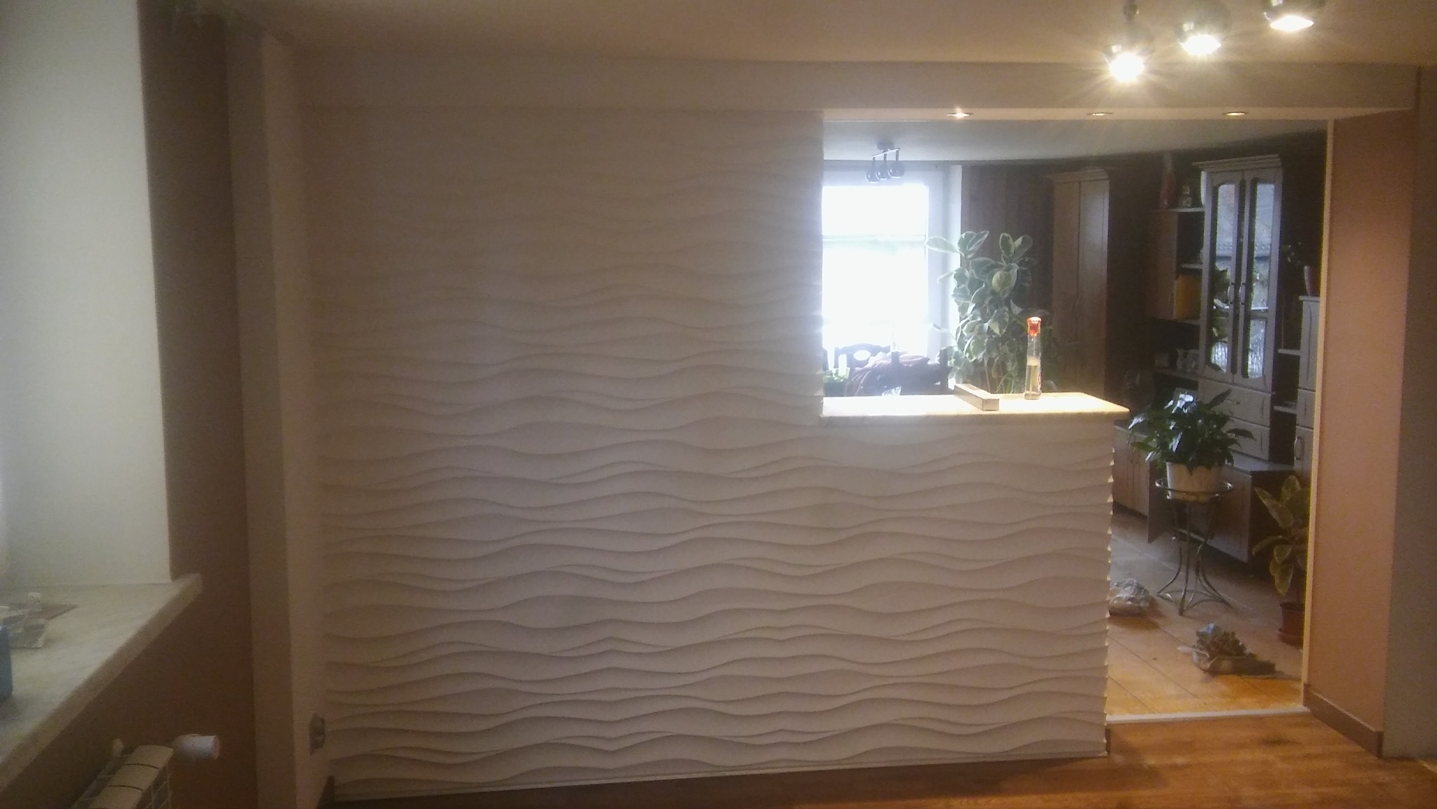 Ściana z dekoracyjną, falistą strukturą 3D, oddzielająca przestrzeń, z widocznym oknem i wyposażeniem wnętrza w tle.